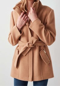 Camelkleurige wollen mix jas met hoge kraag, ceintuur in de taille, grote zakken en een ritsaccent. Zachte textuur, nauwsluitende pasvorm.