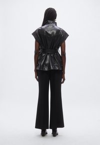 Zwart leren wikkelshirt met een hoge kraag, korte mouwen en een taille met riem, gecombineerd met zwart broek met wijde pijpen en enkellaarsjes met hak.