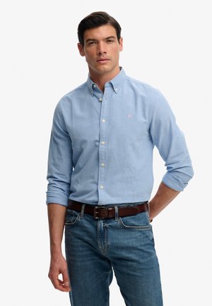 Chemise bleu clair à boutons de coupe classique, dotée d'une texture douce et d'un petit logo rose, associée à un jean en denim bleu et une ceinture marron.