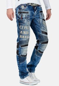 Jeans in denim blu con texture miste, toppe sui ginocchi e dettagli con logo. Più tasche e accenti di cucito arricchiscono il design.