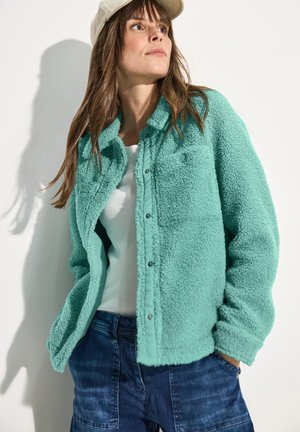 Veste en polaire couleur aquamarine avec un devant à boutons, un col montant, et deux poches, portée par-dessus un haut blanc et assortie à un jeans bleu.
