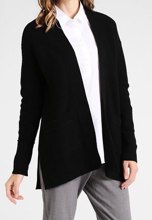 Woman wearing a black open-front cardigan over a white buttoned shirt and gray pants, standing with relaxed arms.