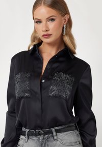 Zwarte satijnen blouse met twee gewatteerde zakken met metalen stiksels, knopen aan de voorkant en lange mouwen. Gecombineerd met lichtgrijze spijkerbroek en een zwarte riem.