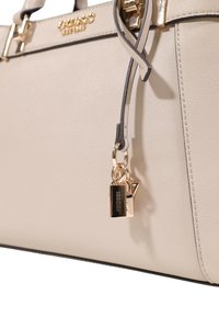 Sac à main en simili cuir beige avec quincaillerie dorée, présentant un charme en forme de serrure et une étiquette décorative. La surface est à la fois texturée et lisse.