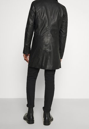 Man draagt een lange zwarte leren jas, zwarte broek en zwarte enkellaarsjes, staand met de rug naar voren tegen een effen achtergrond.