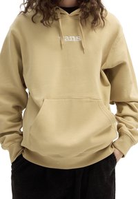 Felpa beige con cappuccio e tasca a marsupio, con ricamo del logo "Vans" bianco sul petto, realizzata in un materiale morbido e spazzolato.
