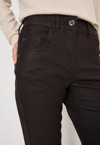 Donkerbruine broek met een gladde textuur, voorzien van een voorzak, gedetailleerde stiksels en een glanzende knop bij de tailleband.