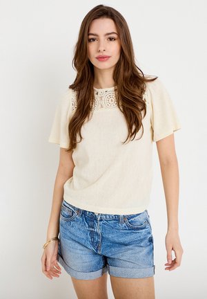 Jonge vrouw met lang bruin haar, gekleed in een crème kanten top en blauwe opgerolde denim shorts, staand tegen een witte achtergrond.