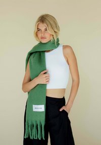 Groene wollen sjaal met franjes aan de randen en een wit label, gedragen rond de hals, gecombineerd met een witte aansluitende tanktop en zwarte broek.