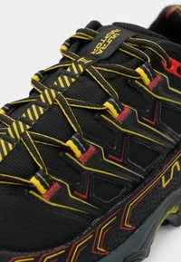 Μαύρο αθλητικό παπούτσι τρεξίματος με υφή στην πάνω πλευρά, με κίτρινες και κόκκινες λεπτομέρειες, ελαστικά κορδόνια και ανθεκτική σόλα με σχέδιο.