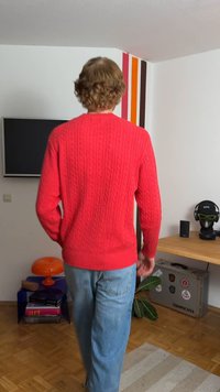 Rød strikket sweater med kabelmønster, der har en ribbet udskæring og kant. Båret med lyseblå jeans. Baggrunden i rummet inkluderer en plante.