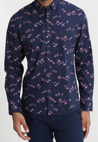 Dunkelblaues Hemd mit Knöpfen und einem Muster aus rosa Vögeln, taillierte Passform, lange Ärmel und ein Kragen. Verfügt über fünf Knöpfe auf der Vorderseite.