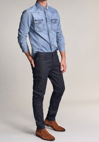 Camicia in denim azzurro con due tasche sul petto e jeans blu scuro. Stivaletti alla caviglia in suede marrone. Outfit casual con design aderente.