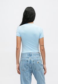 Άτομο με μακριά μαύρα μαλλιά, ντυμένο με ανοιχτό μπλε κοντομάνικο πουκάμισο και ανοιχτό μπλε ριγέ τζιν, να στέκεται με το πρόσωπο προς τα πίσω σε λευκό φόντο.