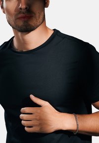 Musta lyhythihainen t-paita, jossa on pyöreä kaula-aukko, henkilö, joka pitää kättään vartalon ympäri. Ranteessa hopeaketju-rannekoru.