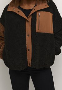 Henkilö, joka pukeutunut mustaan ja ruskeaan fleecetakkiin, jossa on nepparit ja vetoketjullinen rintatasku, yhdistettynä mustiin housuihin.