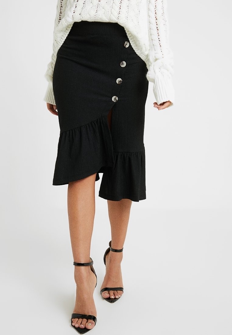 Black denim skirt featuring a ruffled hem, front buttons, and a side slit. Worn with a knitted white sweater and black ankle strap heels.