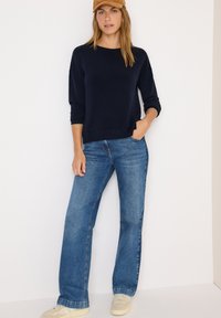 Navyfarbener Pullover mit Dreiviertelärmeln, kombiniert mit hellblauen ausgestellten Jeans und beigen Sneakers, accessoriert mit einer hellbraunen Mütze.