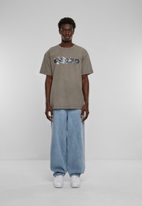 Grå overdimensjonert t-skjorte med et metallisk grafisk design på brystet, kombinert med vide lyseblå denimjeans og hvite joggesko.
