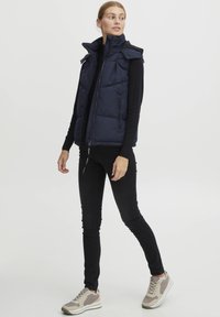 Marinblå pufferväst med dragkedja, hög krage och sidofickor, bärs över en svart långärmad topp, ihop med svarta jeans och grå sneakers.