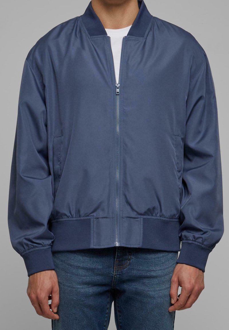 Homme portant une veste bomber bleu marine zippée sur une chemise blanche et un jean bleu, debout avec les mains détendues le long du corps.