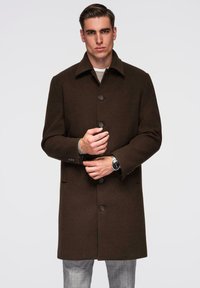 Ruskea villakangastakki, jossa on terävä kaulus, viisi edessä olevaa nappia ja räätälöity istuvuus. Kankaassa on tekstuuri ja sivutaskut, takki on puettu ruudullisten housujen päälle.