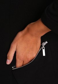 Černá tkanina s kapsou na zip; viditelný kovový zip s obdélníkovým táhlem; ruka položená uvnitř kapsy.