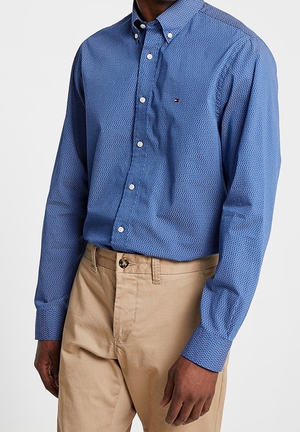 Homme portant une chemise bleue à motifs boutonnée, rentrée dans un pantalon beige avec fermeture par bouton et fermeture éclair, debout, les bras détendus le long du corps.