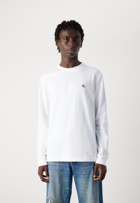 Hvit langermet t-skjorte med rund hals, med en liten svart 'ck' logo på venstre bryst. Kombinert med lyseblå slitte jeans.