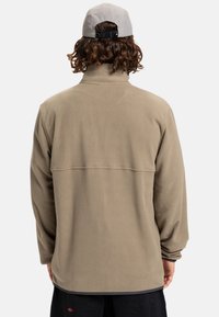 Beige fleece jacket with a high collar, long sleeves, and a horizontal seam across the back. Worn with a light gray cap.