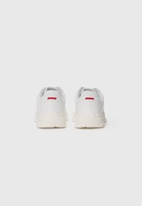 White sneakers with gray accents, featuring a textured mesh upper, thick cream rubber sole, and red logo on the heel.