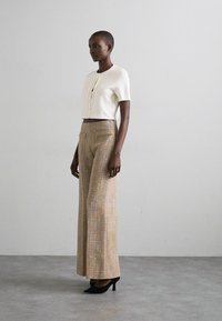Cremefarvet cropped sweater med knapdetalje, parret med højttaljede, brede bukser i tekstureret beige med subtile metalliske tråde. Sorte hæle.