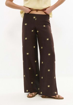 Personne portant un pantalon large foncé avec des motifs jaunes de soleil et des sandales marron, tenant l'ourlet d'un haut jaune clair.