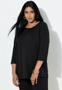 Czarna, miękka bluzka z długim rękawem, zaokrąglonym dekoltem i drobnymi koralikami na wysokości klatki piersiowej, zestawiona z czarnymi spodniami.