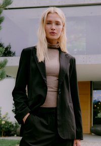 Blonde Frau, die ein schwarzes Blazer und eine schwarze Hose mit einem beigen Rollkragenpullover trägt, steht im Freien in der Nähe eines modernen Gebäudes und grüner Pflanzen.