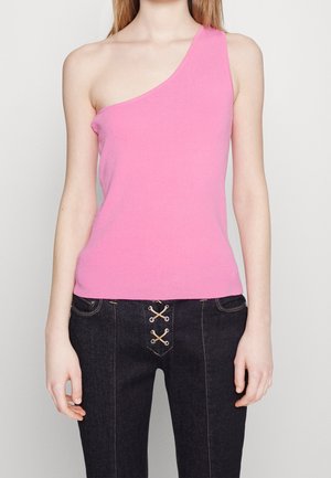 Vrouw draagt een mouwloze, asymmetrische roze top en donkere, hoog getailleerde broek met veterdetail aan de voorkant, staand tegen een effen achtergrond.