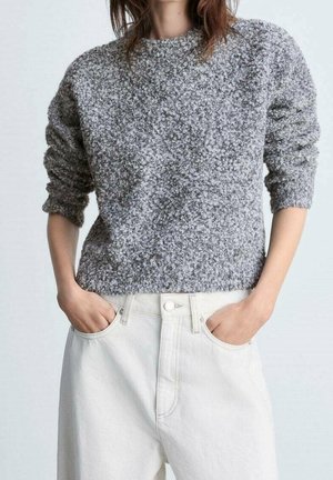 Person trägt einen strukturierten grauen Pullover und hochtaillierte weiße Hosen mit den Händen in den vorderen Taschen.