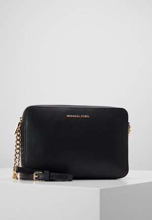 Sac bandoulière Michael Kors en cuir noir avec une chaîne dorée et une fermeture éclair, présenté sur un socle blanc devant un fond uni.