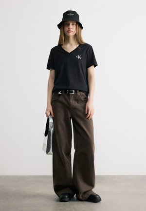 Ung kvinde iført sort Calvin Klein bucket hat og V-udskærings T-shirt, løstsiddende mørkebrune bukser, sorte sko, holder en sølvfarvet tote taske.