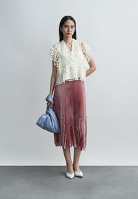 Femme portant un haut en dentelle crème, une jupe midi plissée rose poudré avec des détails floraux, des chaussures blanches à bouts pointus, tenant un sac à main en satin bleu.