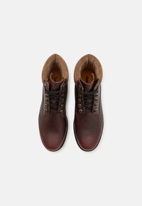 Stivali ankle in pelle marrone con una finitura liscia, collo in suede color cammello a contrasto, lacci neri e occhielli in metallo. Suole in gomma piatta.