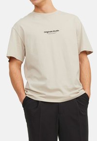 Beige cotton t-shirt with a loose fit, short sleeves, and a black "Originals Studio" graphic printed on the chest.