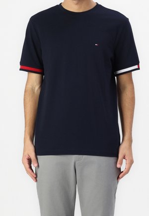 Homme portant un t-shirt bleu marine à manches courtes avec des bordures de manches rayées rouge, blanc et noir et un pantalon gris, debout sur fond blanc.