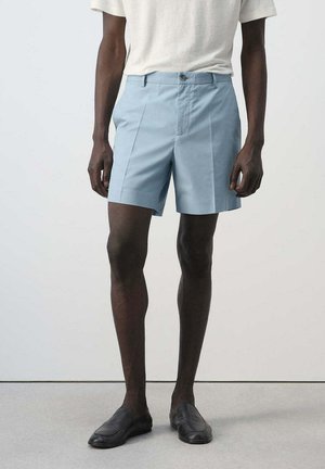 Hombre con pantalones cortos ajustados de color azul claro, camiseta blanca y zapatos negros sin cordones, de pie frente a un fondo liso.
