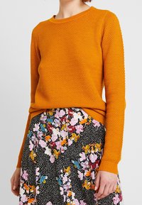 Orange stickad tröja med ett strukturerat mönster, rundad halsringning och ribbade muddar, matchad med en blommig svart kjol med färgglada accenter.