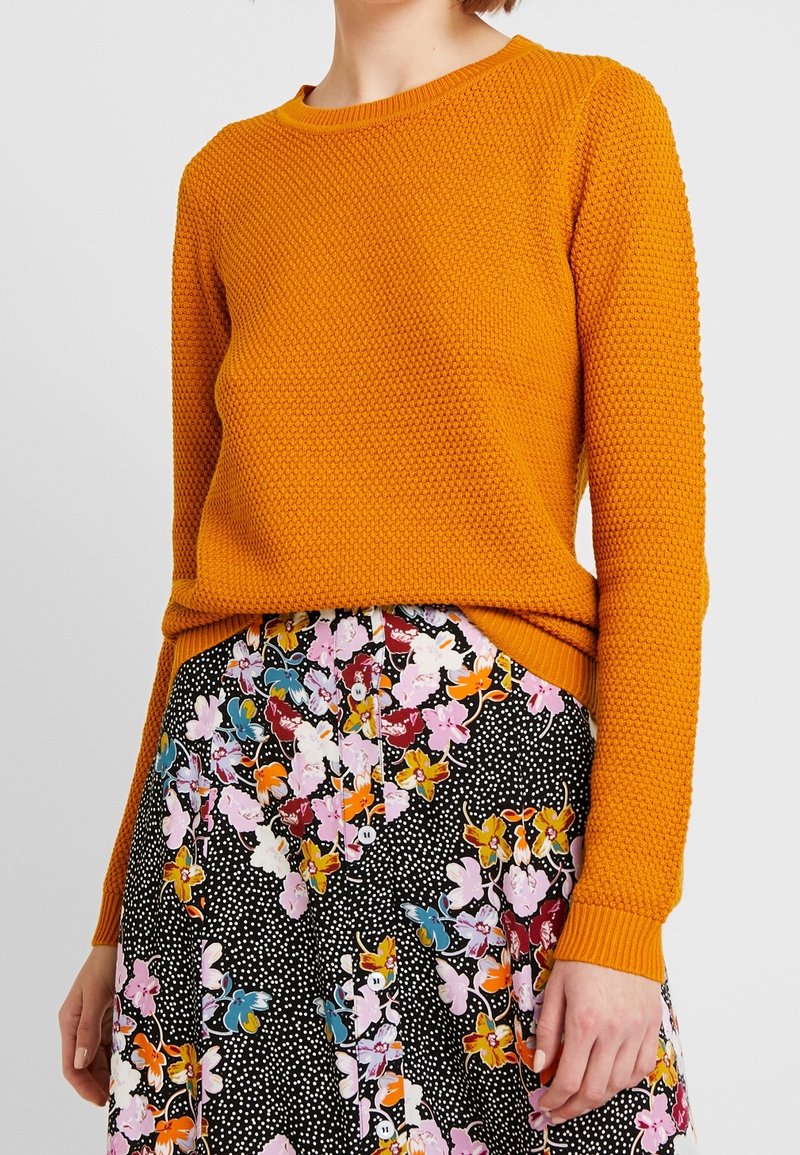 Orange stickad tröja med ett strukturerat mönster, rundad halsringning och ribbade muddar, matchad med en blommig svart kjol med färgglada accenter.