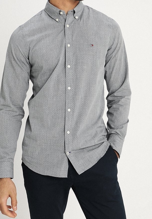 Homme portant une chemise grise ajustée à motifs, boutonnée avec col fermé et un pantalon foncé, une main dans la poche, debout devant un fond uni.
