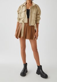 Beżowa krótka kurtka z dwiema kieszeniami na piersi, zestawiona z brązową plisowaną spódnicą. Czarne masywne botki za kostkę z gumowymi podeszwami.