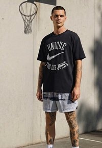 Tetovált férfi fekete "Unique Tous Les Jours" feliratú pólóban és világos sortban áll egy szabadtéri kosárlabdapályán a gyűrű közelében.