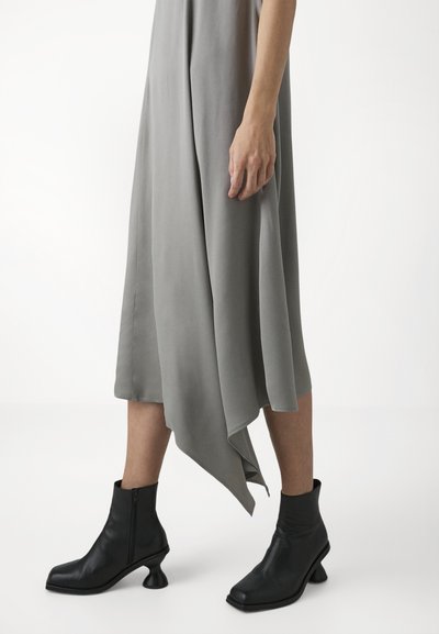 Grijze midi-jurk met een asymmetrische zoom, gecombineerd met zwarte leren enkellaarsjes met een opvallende gebogen hak en ritssluiting.